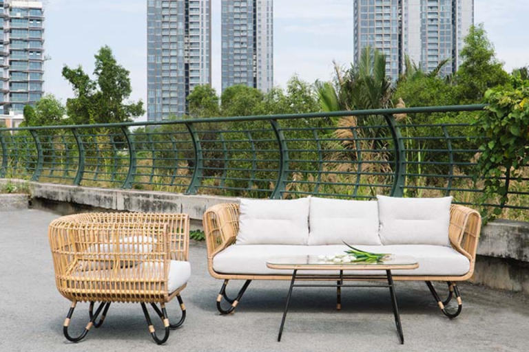 Bộ bàn ghế mây nhựa Alozo gồm sofa 2 chỗ, ghế đơn và bàn trà, đặt ngoài trời trên ban công với nền cây xanh và tòa nhà phía sau.