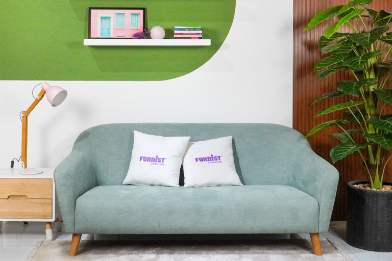 Không gian phòng khách hiện đại với ghế sofa màu xanh mint, hai gối tựa lưng màu trắng in logo Furnist. Bên cạnh là bàn gỗ nhỏ có đèn bàn màu hồng nhạt, phía sau là tường trang trí mảng xanh lá cây và một chậu cây cảnh lớn.